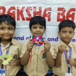 Three smiling young boys in beige school uniforms hold colorful crafts at a banner backdrop for a school event